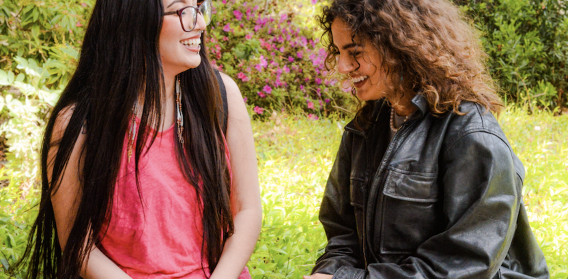 two students talking and laughing while sitting on wall with green space in the background